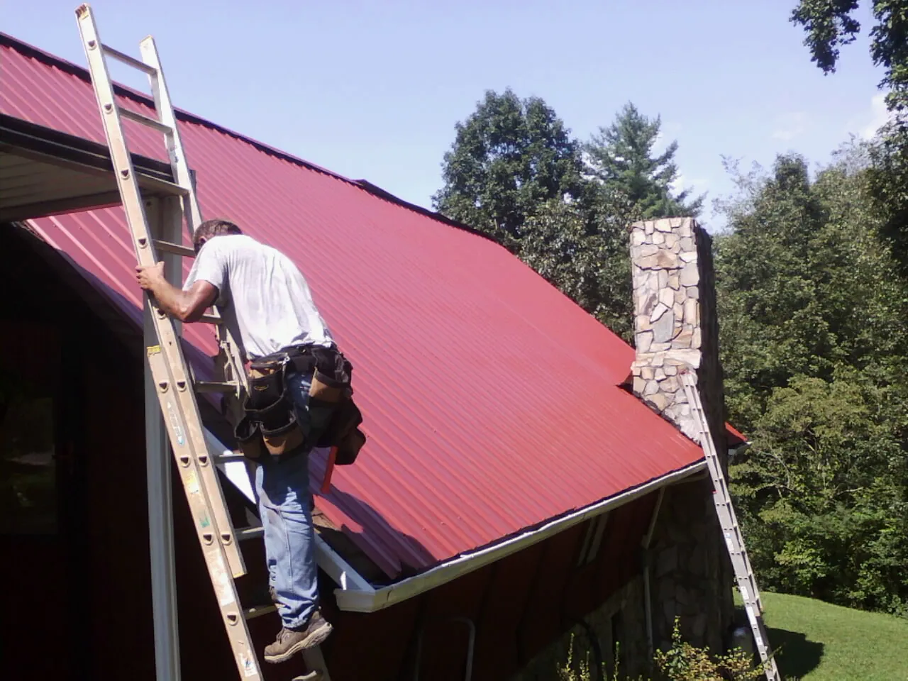 Roofer on a ladder beside a red metal roof and stone chimney — Soffit & Fascia Repair by Roofing Brothers in Bly